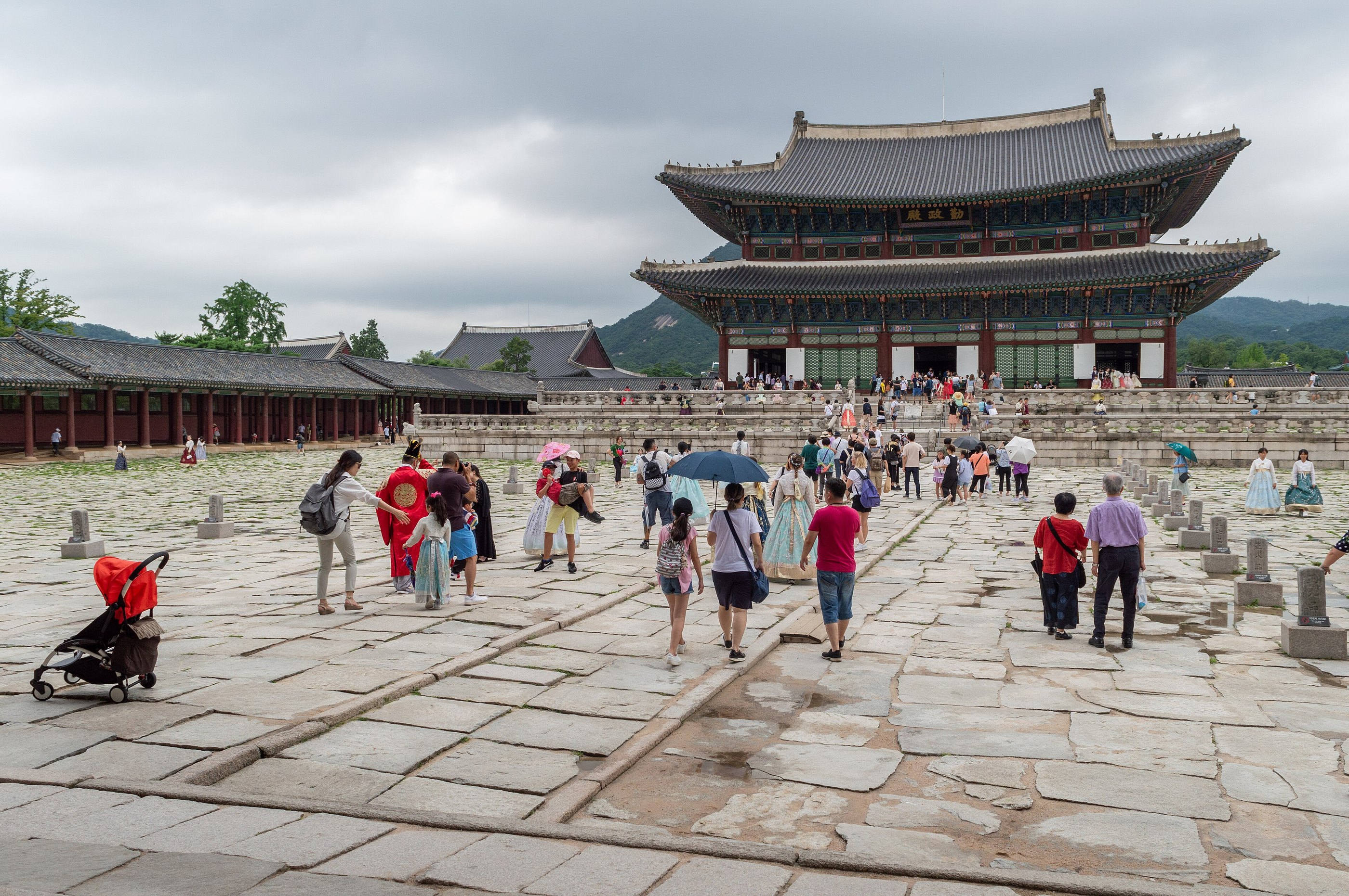 The throne hall of Gyeongbokgung across its wide flagstone courtyard, wooded hills behind it, with visitors walking the stone path and several of them in hanbok
